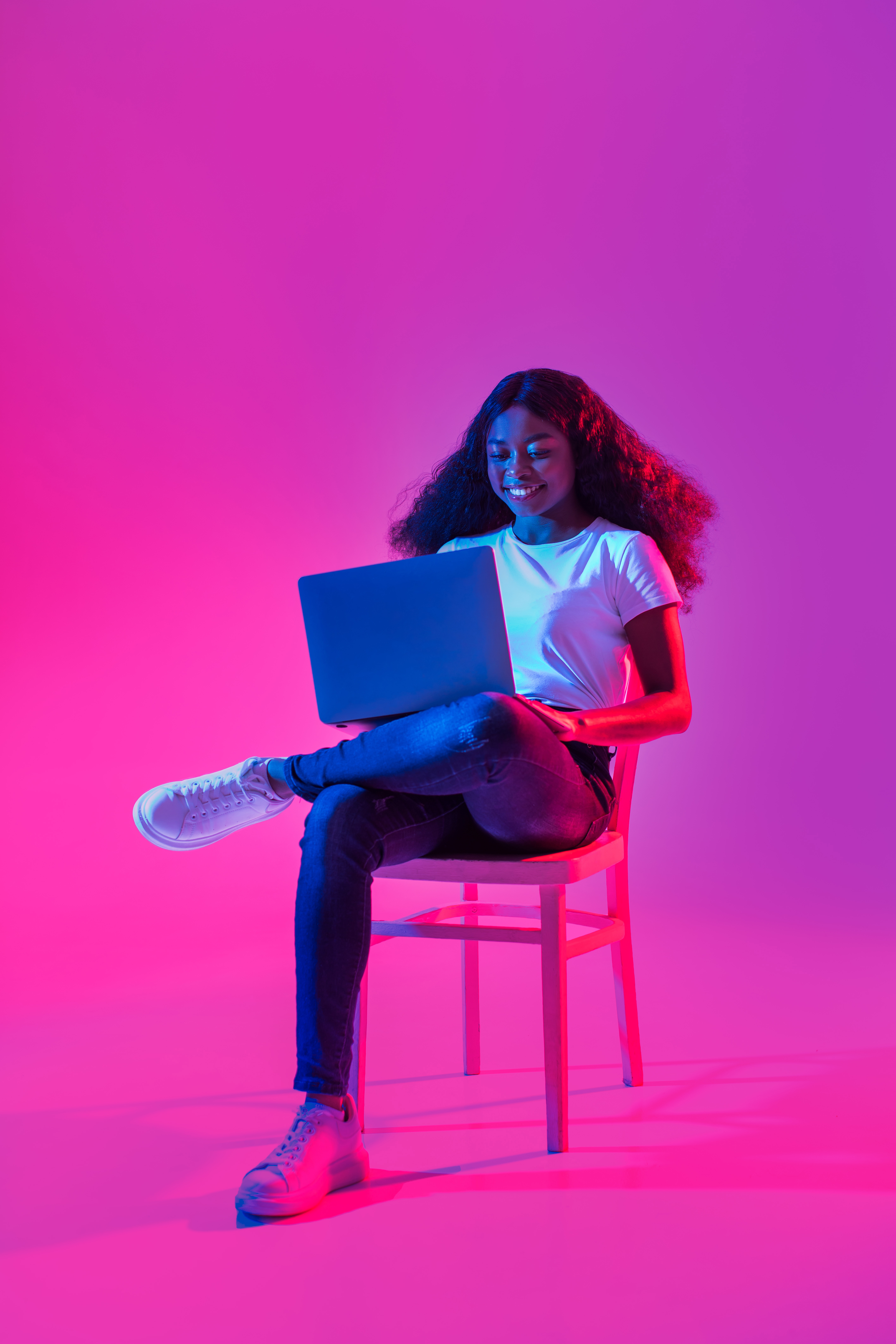 Woman sat on a chair with neon backlighting, looking at a laptop, smiling
